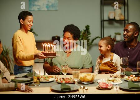 Portrait d'un adolescent souriant apportant un gâteau d'anniversaire à une heureuse grand-mère afro-américaine célébrant avec sa famille Banque D'Images