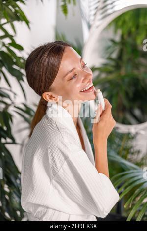 Portrait d'une jeune femme mignonne avec une belle peau souriante en appliquant un baume à lèvres sur ses lèvres, debout dans la salle de bains Banque D'Images