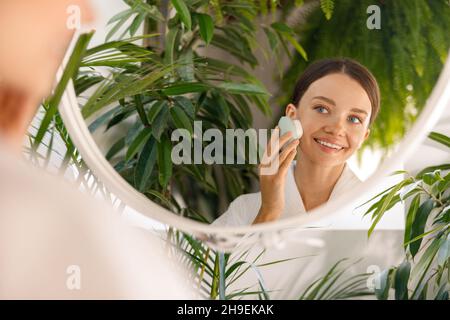 Portrait de la jeune femme se regardant joyeusement dans le miroir tout en nettoyant sa peau à l'aide de la brosse silicone visage dans la salle de bains décorée avec Banque D'Images