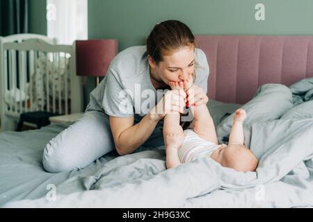 Une jeune maman heureuse à tête rouge s'amuse, embrassant les petits pieds d'un nouveau-né allongé sur le lit. Banque D'Images