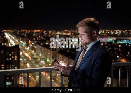 Jeune homme d'affaires prospère avec messagerie texte smartphone tout en se tenant sur le balcon contre les veilleuses Banque D'Images
