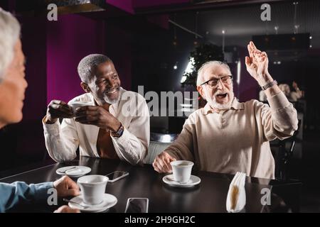 un homme de premier plan émerveillé en regardant loin et en agitant la main tout en passant du temps avec des amis multiethniques dans un café Banque D'Images