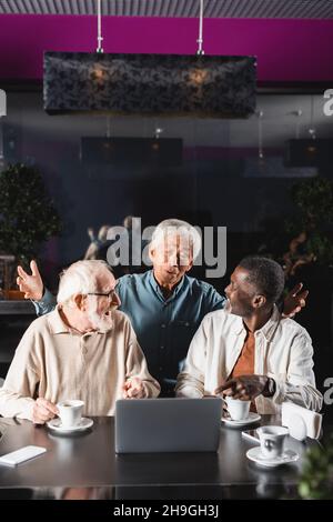 un homme asiatique émerveillé montrant un geste de wow près d'amis interraciaux confirmés pointant vers un ordinateur portable dans un café Banque D'Images