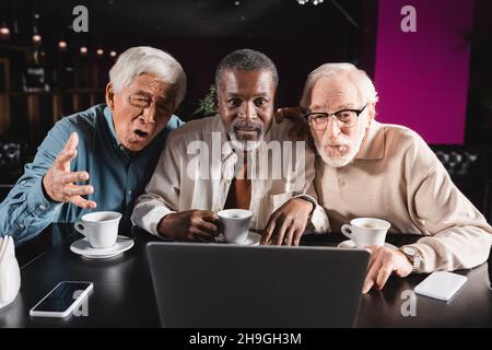 un homme asiatique stupéfait de pointer de la main près d'amis multiethniques confirmés qui ont un chat vidéo sur ordinateur portable Banque D'Images