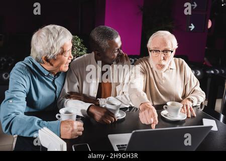 un homme de grand âge émerveillé se dirigeant vers un ordinateur portable près d'amis multiethniques souriants dans un café Banque D'Images