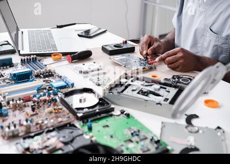 Les mains d'un jeune homme africain réparant la carte de circuit imprimé avec un fer à souder tout en étant assis près d'un bureau devant un ordinateur portable Banque D'Images