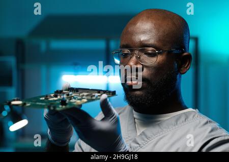 Sérieux réparateur africain en gants et uniforme tenant la carte de circuit imprimé et le regardant en laboratoire Banque D'Images