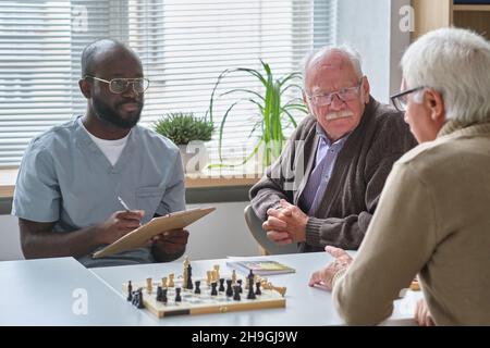 Un médecin africain prend des notes en carte médicale tout en parlant à un homme âgé pendant son jeu d'échecs avec un ami à la table Banque D'Images