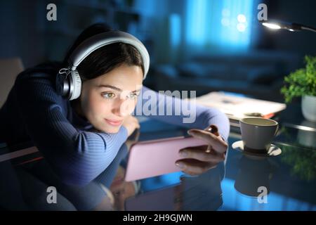 Femme détendue, portant un casque, regardant des médias sur un smartphone la nuit à la maison Banque D'Images