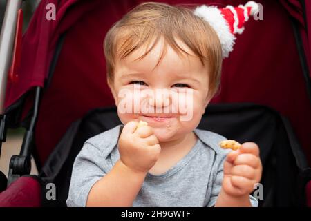 bébé dans le petit chapeau de père noël est assis dans la poussette, sourit, mange un bagel Banque D'Images