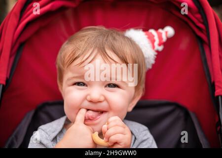 bébé est assis dans la poussette rouge. l'enfant mange un bagel et montre sa langue. Banque D'Images