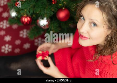 Une belle jeune femme dans un chandail rouge tient une boîte avec un cadeau attaché avec un ruban rouge à la maison près d'un arbre de Noël sur une couverture chaude.Foc sélectif Banque D'Images