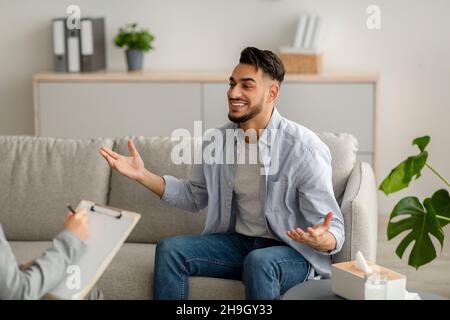 Jeune homme arabe heureux parlant à son psychologue, recevant de l'aide professionnelle, assis sur un canapé à la clinique de santé mentale Banque D'Images