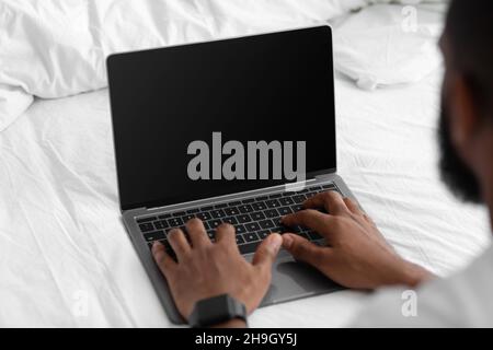 Un jeune afro-américain regarde un ordinateur portable avec écran blanc, travaille à distance sur un lit blanc dans la chambre Banque D'Images