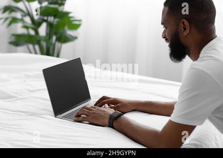 Homme barbu afro-américain souriant du millénaire regarde l'ordinateur portable avec écran blanc, travaille à distance sur un lit blanc Banque D'Images