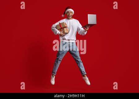 Un homme dans un chapeau de père Noël avec un ordinateur portable et des cadeaux qui sautent sur un fond rouge Banque D'Images
