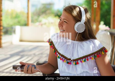 Portrait d'une jolie fille assise sur le canapé avec un casque et un téléphone portable dans ses mains.Une jeune fille écoute de la musique tout en passant du temps à l'extérieur lors d'une journée ensoleillée d'été.Copier l'espace. Banque D'Images