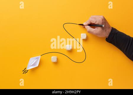 Main masculine dessine une ligne courbe qui guide un bateau en papier pour traverser les barrières.Orientation, assistance, leadership ou pour surmonter les obstacles concept. Banque D'Images