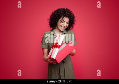 Vue avant portrait d'une femme portant une robe, ouvrant une boîte cadeau, regardant à l'intérieur avec l'expression heureuse isolée sur fond rose.Poil bouclés Banque D'Images