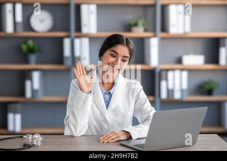 Sympathique gai jeune femme indienne médecin en manteau blanc travaille sur le lieu de travail et fait des vagues de main à ordinateur portable dans la clinique Banque D'Images