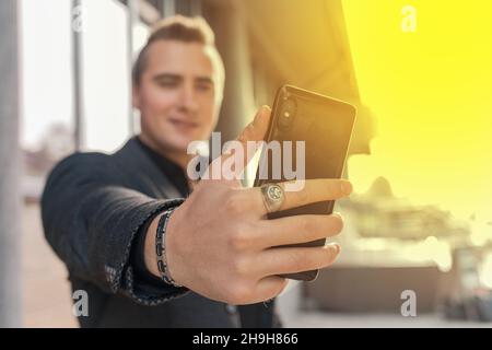 Un jeune homme satisfait tient un smartphone ou un téléphone portable dans sa main, prend un selfie ou parle sur vidéo dans la rue extérieure, en gros plan. Banque D'Images