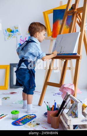 Mignon, sérieux et concentré, blanc garçon de trois ans en chemise bleue et tablier de Jean dessin sur toile debout sur le chevalet.Concept de la petite enfance Banque D'Images