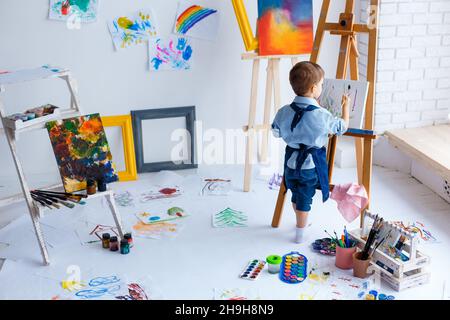 Mignon, sérieux et concentré, blanc garçon de trois ans en chemise bleue et tablier de Jean dessin sur toile debout sur le chevalet.Concept de la petite enfance Banque D'Images