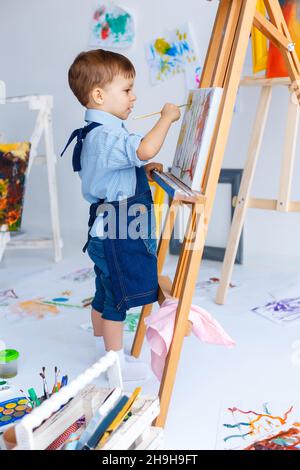 Mignon, sérieux et concentré, blanc garçon de trois ans en chemise bleue et tablier de Jean dessin sur toile debout sur le chevalet.Concept de la petite enfance Banque D'Images