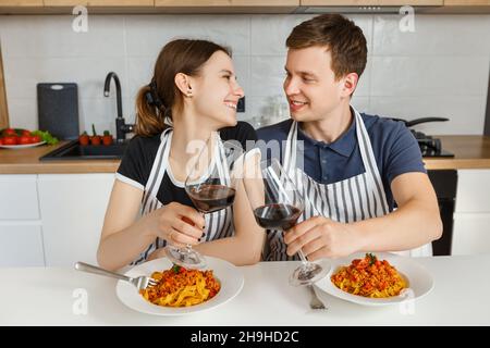 Un couple heureux en tabliers mangeant des pâtes bolognaises avec du vin dans une cuisine moderne.Dîner romantique à la maison.Concept de style de vie domestique, mariage heureux et Banque D'Images