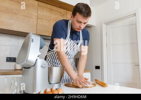 Jeune homme beau en tablier pétriant la pâte dans la cuisine moderne.Concept de boulangerie maison, cuisine masculine et style de vie domestique.Pâtisserie du chef Banque D'Images
