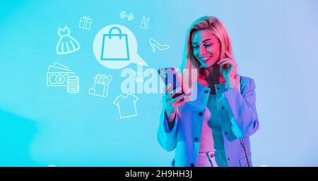 Belle femme souriante dans des vêtements à la mode tenant un smartphone et magasiner en ligne avec des icônes de shopping sur fond bleu.Une fille fait des achats Banque D'Images