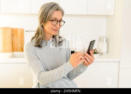 Une femme mûre souriante aux cheveux gris tient le smartphone assis dans la cuisine.Une charmante dame passe du temps libre sur le Web, magasiner en ligne, faire défiler les nouvelles sur le téléphone à la maison Banque D'Images