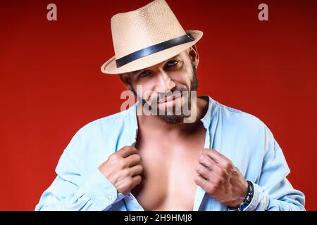 Cuban ou Latino avec le corps tanné dans le chapeau et la chemise pose sur fond rouge.Studio portrait d'un homme barbu de 30-40 ans.Un homme brutal regarde l'appareil photo et sourit.Vacances d'été. Banque D'Images