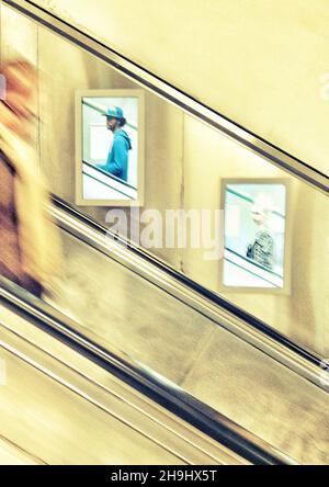 Une personne descendant un esclator sur le métro de Londres avec deux écrans de publicité récemment installés en arrière-plan (partie d'une série d'images expérimentales prises et traitées sur l'iPhone) Banque D'Images