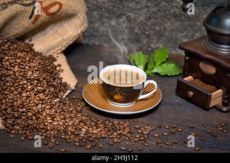 Kaffee Komposition mit dampwender Kaffeetasse, Kaffeebohnen in Kaffeesack, historischer Kaffeemühle und Kaffeeblätter auf einem Holztisch Banque D'Images