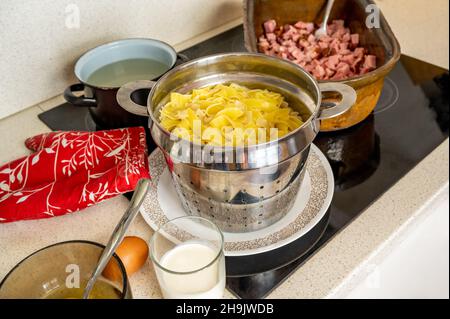 Passoire avec pâtes larges bouillies, œuf, lait, pouleuse rouge, plaque de cuisson en faïence avec viande fumée coupée en cube sur table de cuisson en céramique, repas de préparation. Banque D'Images