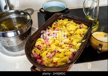 Pâtes larges bouillies avec viande fumée dans une plaque de cuisson en faïence, un support et une casserole sur une table de cuisson en céramique, en gros plan.Préparation du repas. Banque D'Images