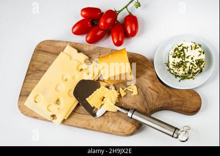 Différentes sortes de fromage (tranché, dur et blanc avec des herbes) sur le plan de cuisine en bois, trancheuse au fromage et tas de tomate sur table blanche, gros plan. Banque D'Images