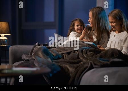 Jolie jeune femme et deux jolies filles assis ensemble sur un canapé confortable, souriant et livre de lecture.La mère passe le soir avec ses filles avant d'aller se coucher. Banque D'Images