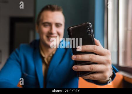 Gros plan, homme d'affaires tient un smartphone ou un téléphone portable noir dans sa main et prend un selfie ou parle sur une liaison vidéo dans un café. Banque D'Images