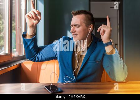 Un beau et drôle homme d'affaires dansant d'apparence européenne élégant portrait, tient un téléphone dans sa main et écoute de la musique dans des écouteurs assis à Banque D'Images