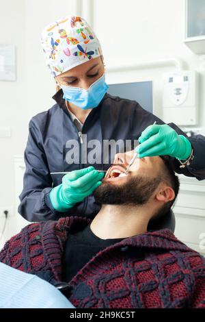 Le dentiste féminin examine un homme patient dans un cabinet dentaire à l'aide d'outils professionnels et d'équipements de protection individuelle. Banque D'Images