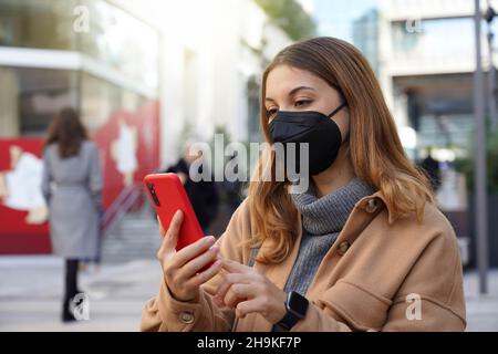 Belle jeune femme avec masque de protection noir FFP2 KN95 utiliser le téléphone pour faire du shopping en ligne dans la rue de ville avec des gens flous sur l'arrière-plan Banque D'Images