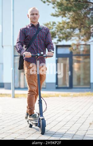 Jeune homme dans un pantalon décontracté et une chemise à cheval sur son scooter électrique, vue de face, bâtiments à l'arrière-plan Banque D'Images