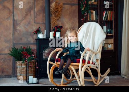 Une jeune fille blonde de trois ans vêque d'une robe verte balance sur un fauteuil à bascule dans une pièce décorée pour Noël.Chambre avec intérieur festif.Un enfant heureux apprécie les vacances d'hiver et l'ambiance de noël. Banque D'Images