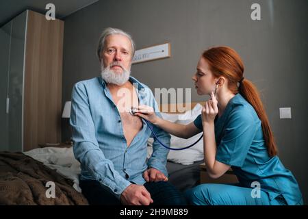 Infirmière féminine ciblée tenant un stéthoscope à l'écoute des poumons ancien patient masculin senior assis sur le lit à la maison. Banque D'Images