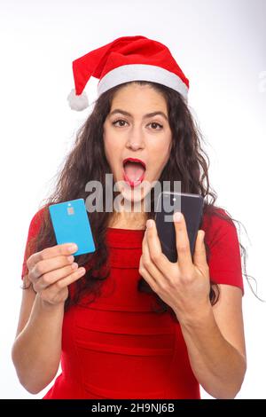 Femme vêtue de robe rouge et chapeau de père Noël tenant une carte de crédit et un mobile avec un geste d'étonnement.Concept d'achat en ligne Banque D'Images