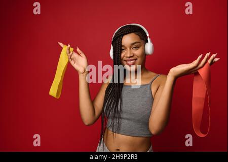 Magnifique femme avec des dreadlocks afro et des écouteurs sans fil tient les bandes élastiques colorées de forme physique dans les mains, souriant avec un sourire joyeux regardant la caméra Banque D'Images
