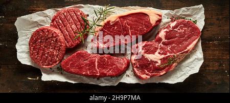 Vue de dessus des sirloins de bœuf cru et des steaks de ribeye avec des côtelettes de hamburger et des brindilles de romarin sur du papier froissé sur une table en bois Banque D'Images