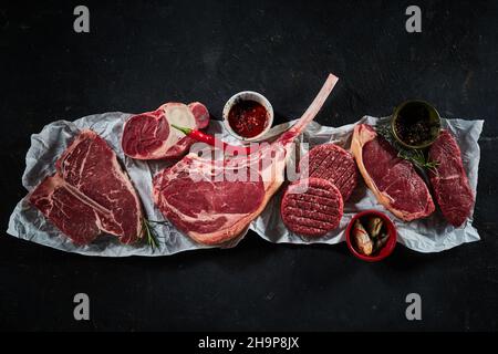 Vue de dessus d'un assortiment de steaks de bœuf cru, y compris le sirloin de tomahawk, servi avec différentes épices et des brindilles de piment et de romarin Banque D'Images